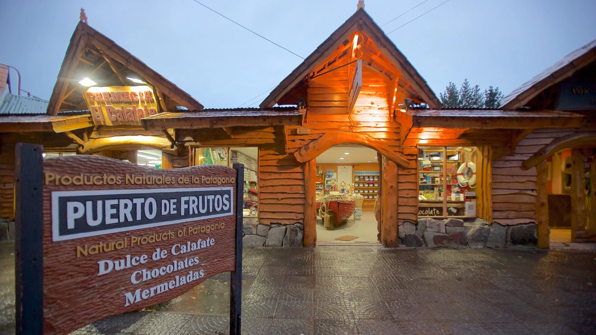 A wooden storefront with a sign that reads 'PUERTO DE FRUTOS' and smaller signs advertising natural products from Patagonia, including Calafate, chocolates, jams, and marmalade, with the interior visible through the entrance.