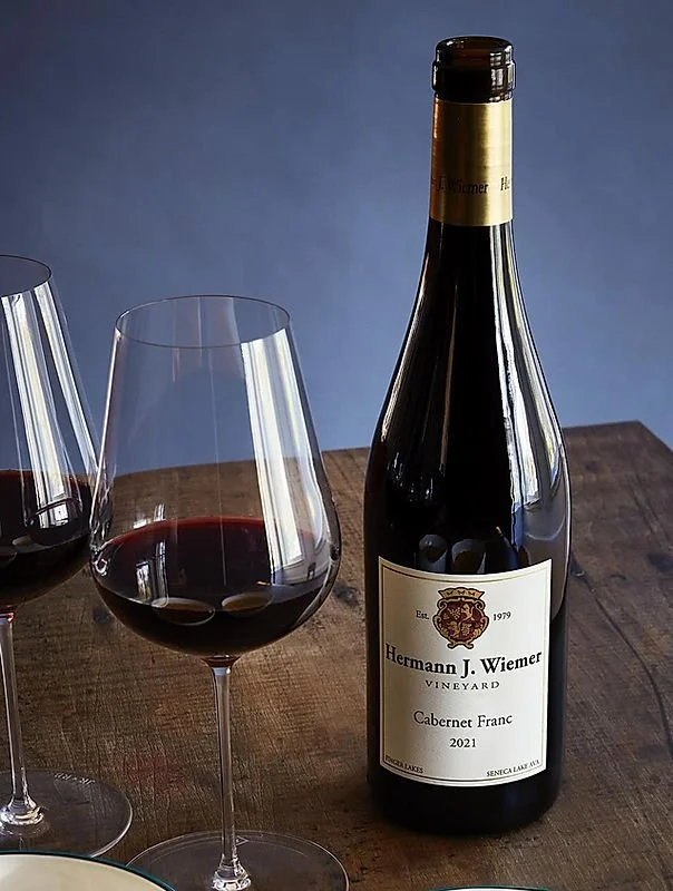 A bottle of Hermann J. Wiemer Vineyard Cabernet Franc 2021 and two glasses of red wine on a wooden table.