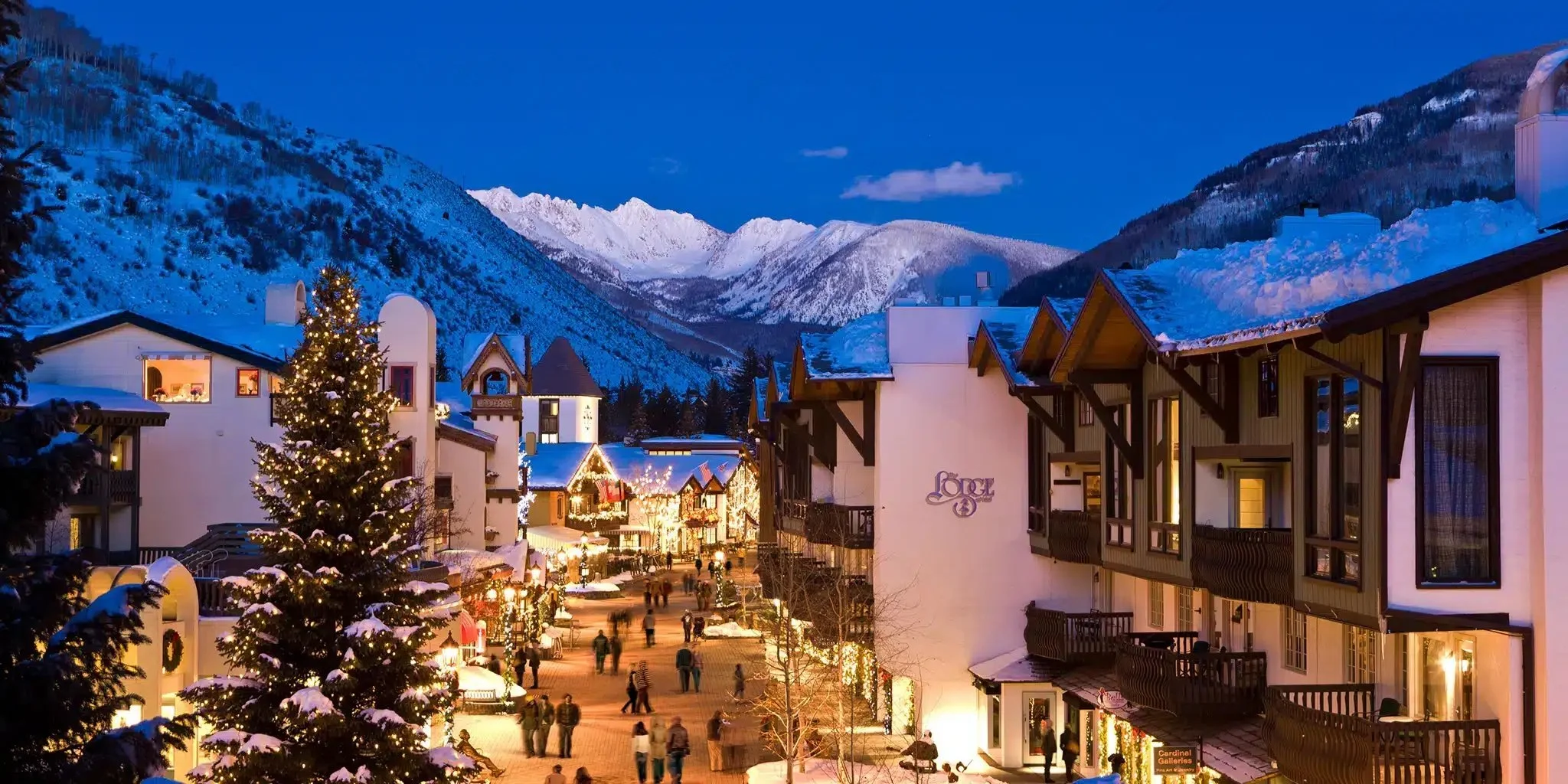 A snow-covered mountain town decorated with Christmas lights and a large Christmas tree, during evening.