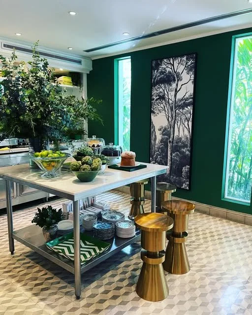 A kitchen with a marble island counter holding bowls of fruit and vegetables. There are three metallic and wooden stools around the island, green walls with large windows, and a black and white forest painting on the wall.