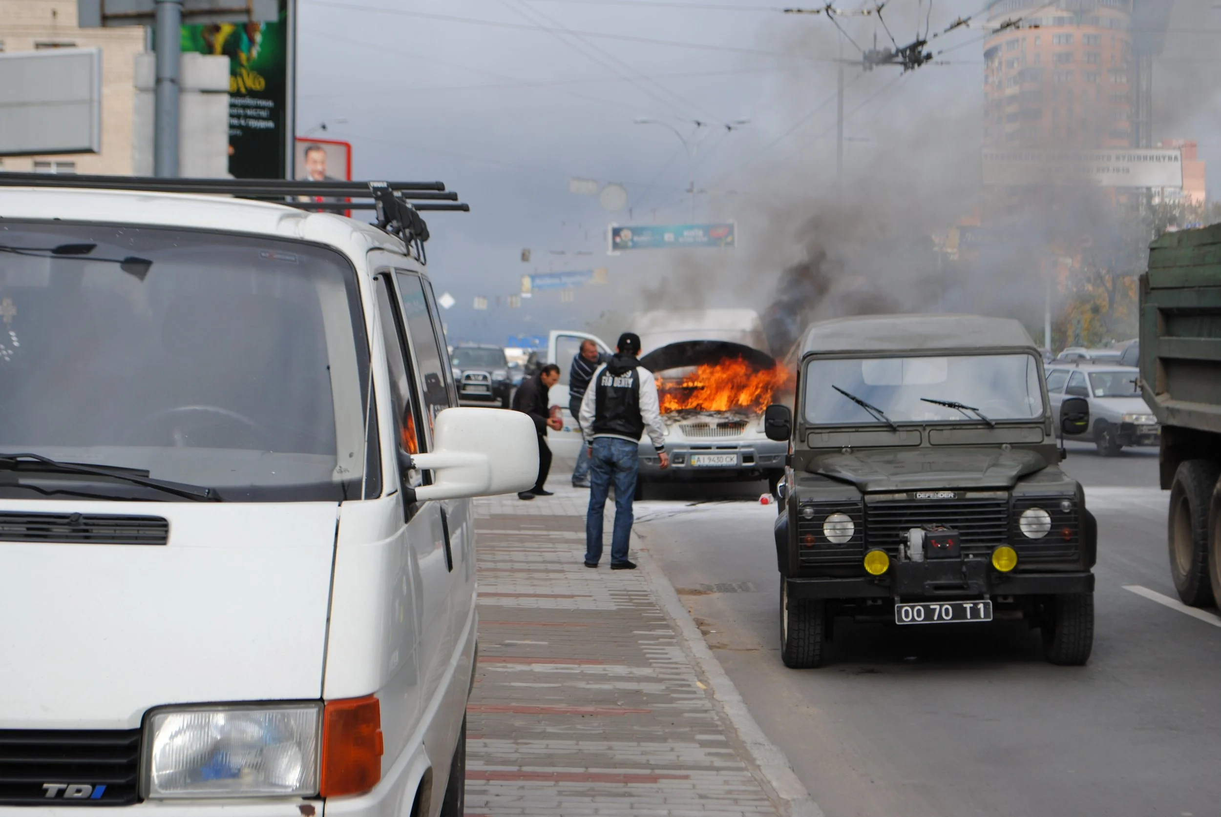 A car on fire on a city street with smoke rising from the engine and several people nearby. Vehicles are parked along the street, and buildings are visible in the background. Kiev November 2011, Marina Parrish ©