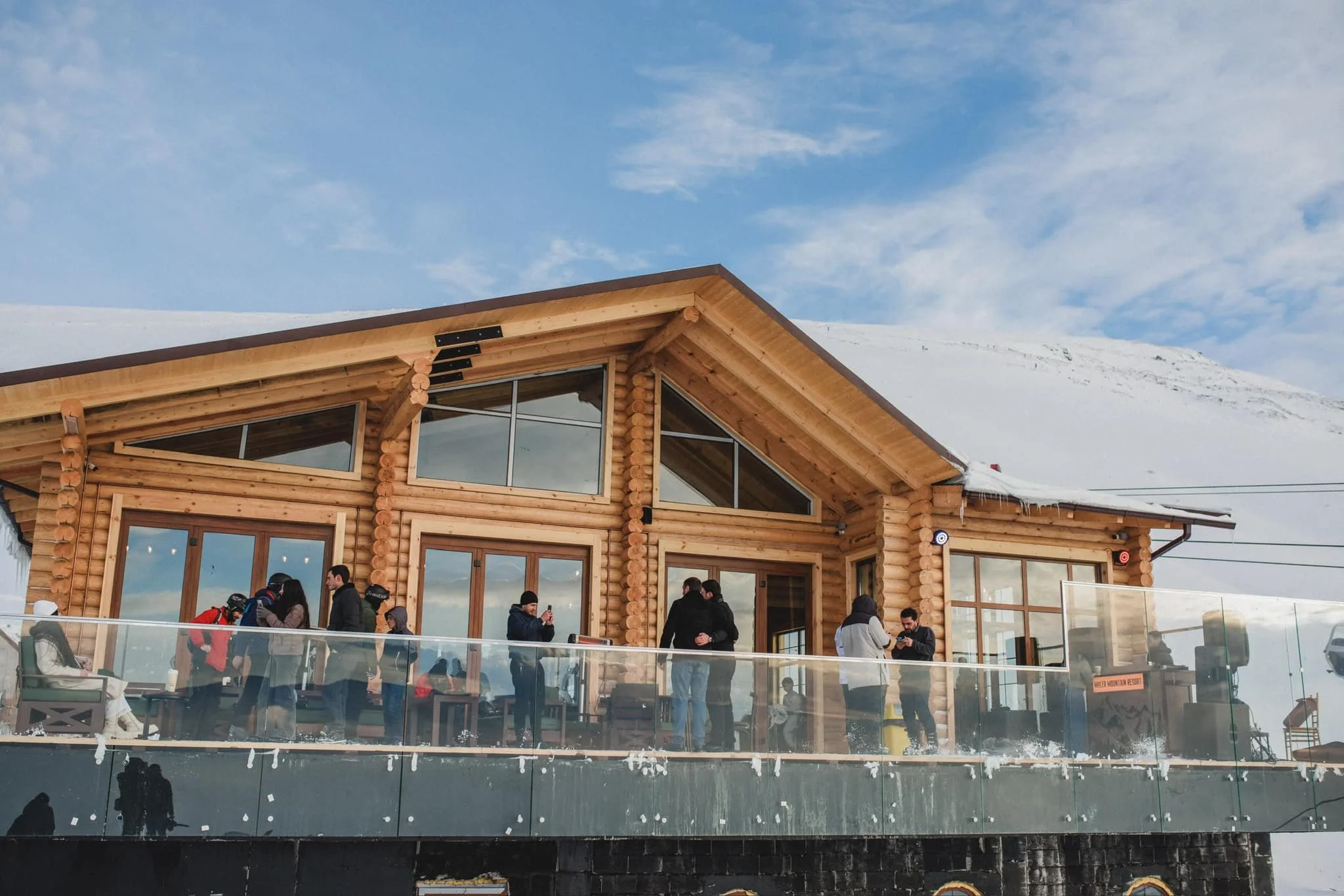 People gathered on a snow-covered wooden deck of a log-style building with large windows, set against a backdrop of snow and blue sky.