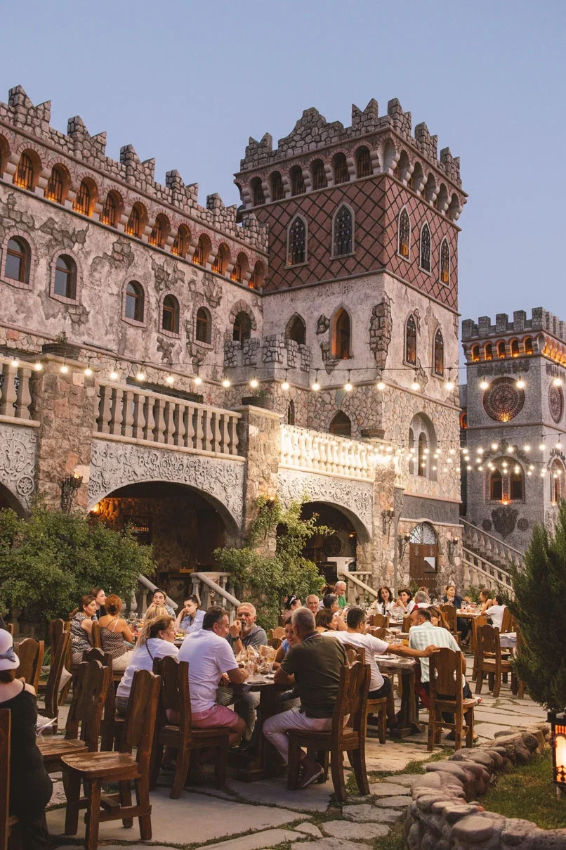 People dining outdoors at a castle-themed restaurant with stone walls, turrets, and string lights in the evening.