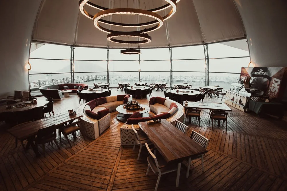 Interior of a modern restaurant or lounge with wooden floors, round tables, and a curved seating area with colorful cushions near large glass windows, overlooking an urban landscape. Circular LED lights hang from the ceiling.