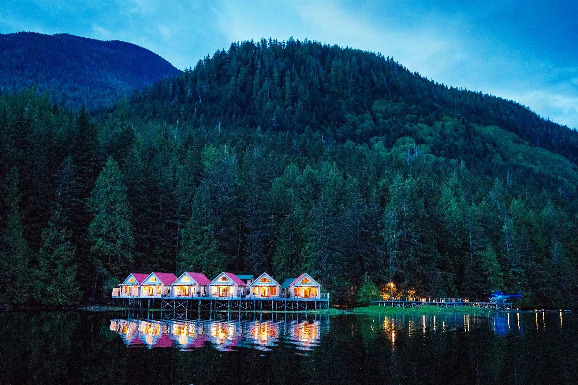 Lakeside view of six illuminated cabins on stilts with a forested mountainside in the background during twilight.