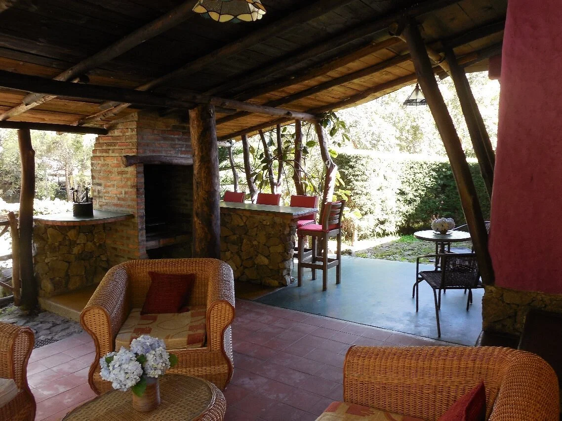 Outdoor patio area with wicker chairs, a brick and stone fireplace, a bar with red chairs, and a small round table with chairs, surrounded by greenery.