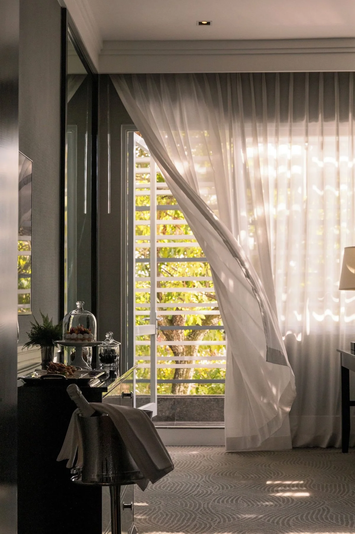 A hotel room with open balcony doors, white curtains, and a view of trees outside. There is a table with desserts and a lamp inside the room.