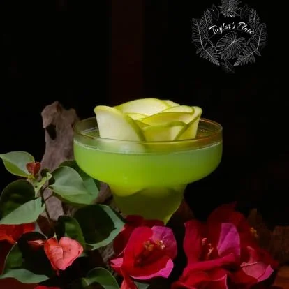 A green cocktail glass filled with a layered green and yellow drink, garnished with fruit slices, surrounded by pink and red flowers and green leaves.