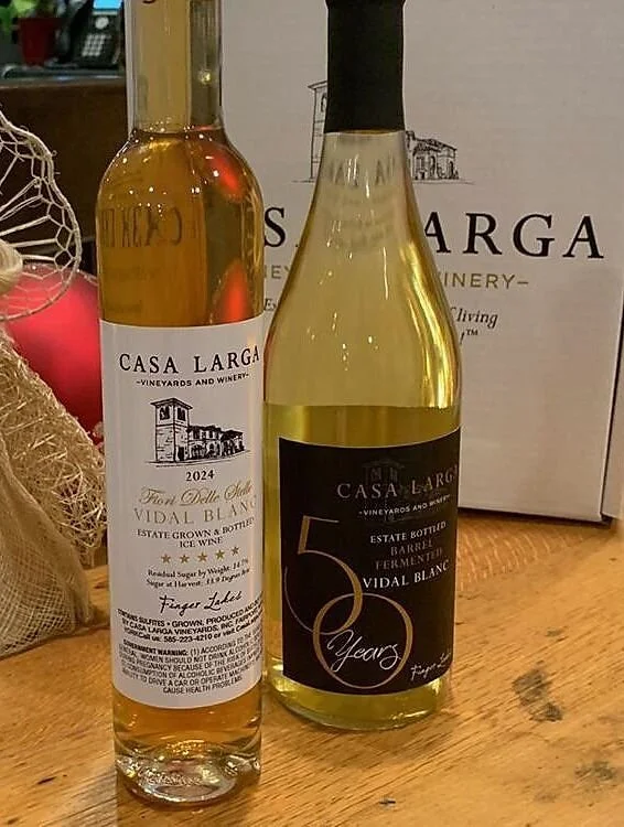 Two bottles of Casa Larga Vidal Blanc white wine on a wooden table, with a white box and decorative items in the background.