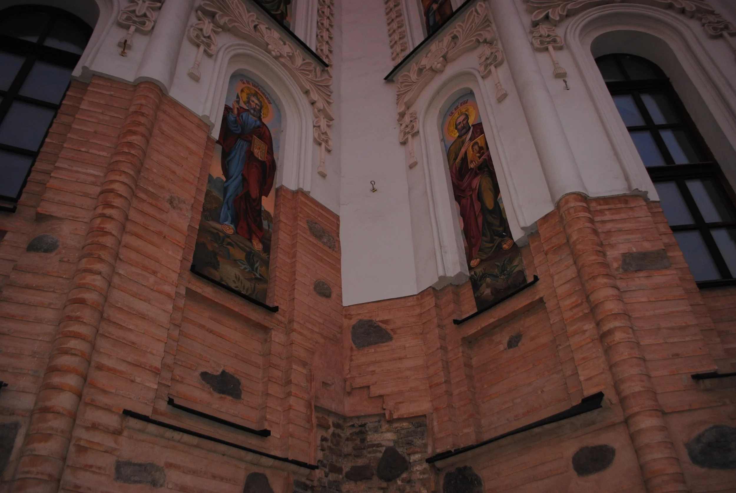 Interior view of a church or cathedral with colorful religious stained glass windows depicting saints, intricate plasterwork, and exposed brick and stone walls.