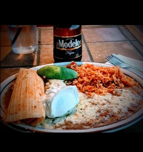 Plate of Mexican food with rice, refried beans topped with cheese, guacamole, sour cream, a tamale, and a fried egg, with a bottle of Modelo Negra beer and a glass of water in the background.