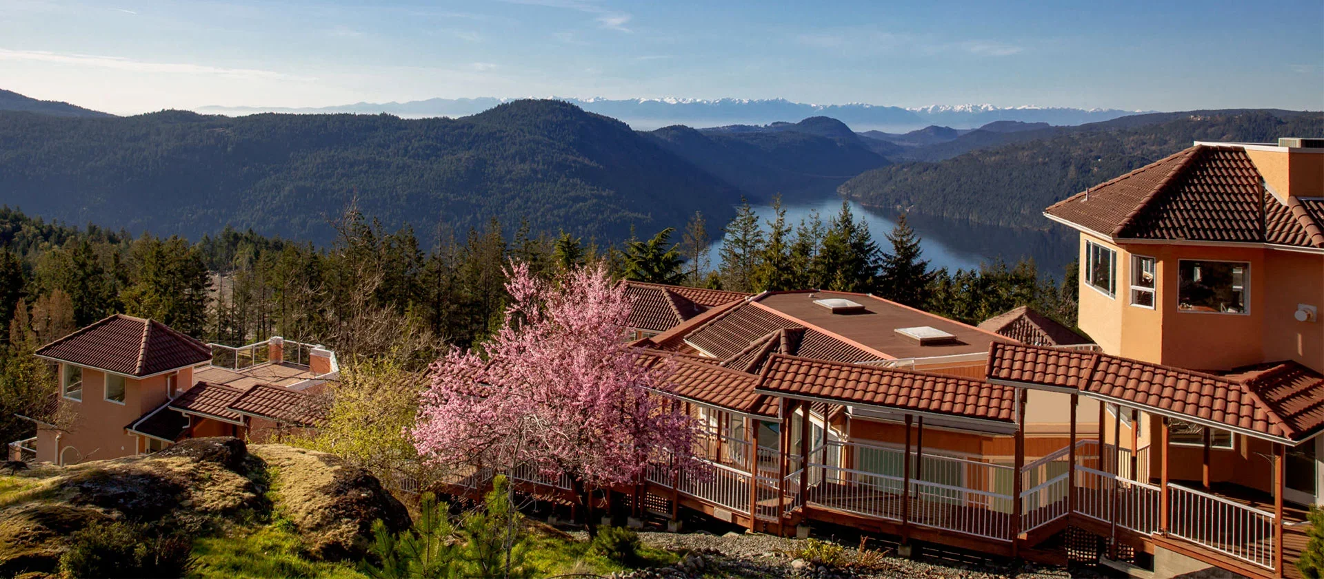A hillside view of houses with red-tiled roofs and a blooming pink tree in front, overlooking a river and forested mountains under a blue sky.
