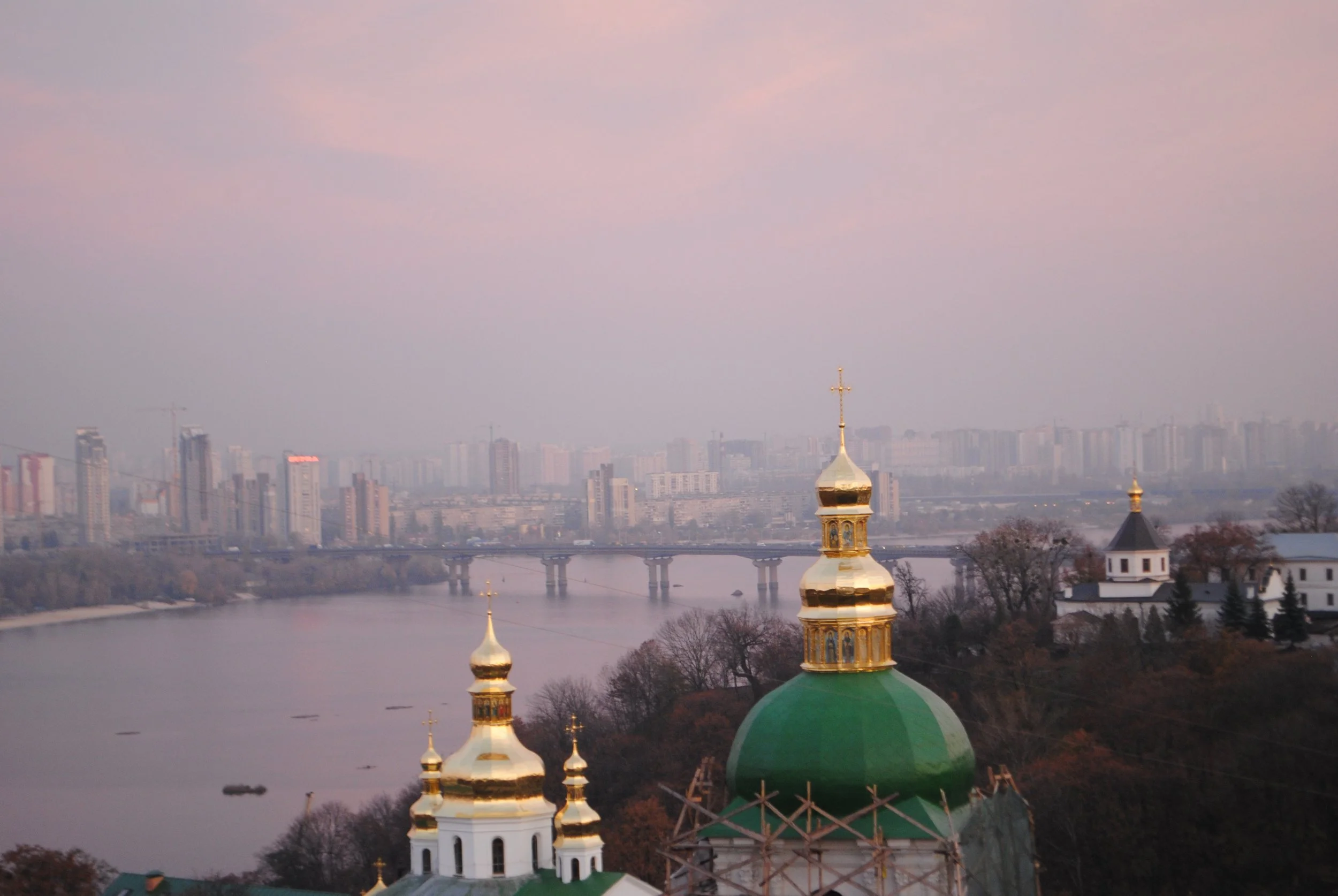 A cityscape with a river in the foreground, a city bridge, and a skyline of tall buildings in the distance under a pink and purple sky. In the foreground, there is an Orthodox church with golden domes and crosses.