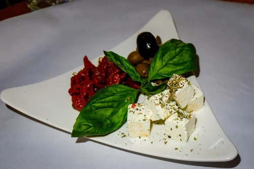 A white triangular plate with slices of feta cheese sprinkled with herbs, fresh basil leaves, sun-dried tomatoes, and green olives on a light-colored tablecloth.