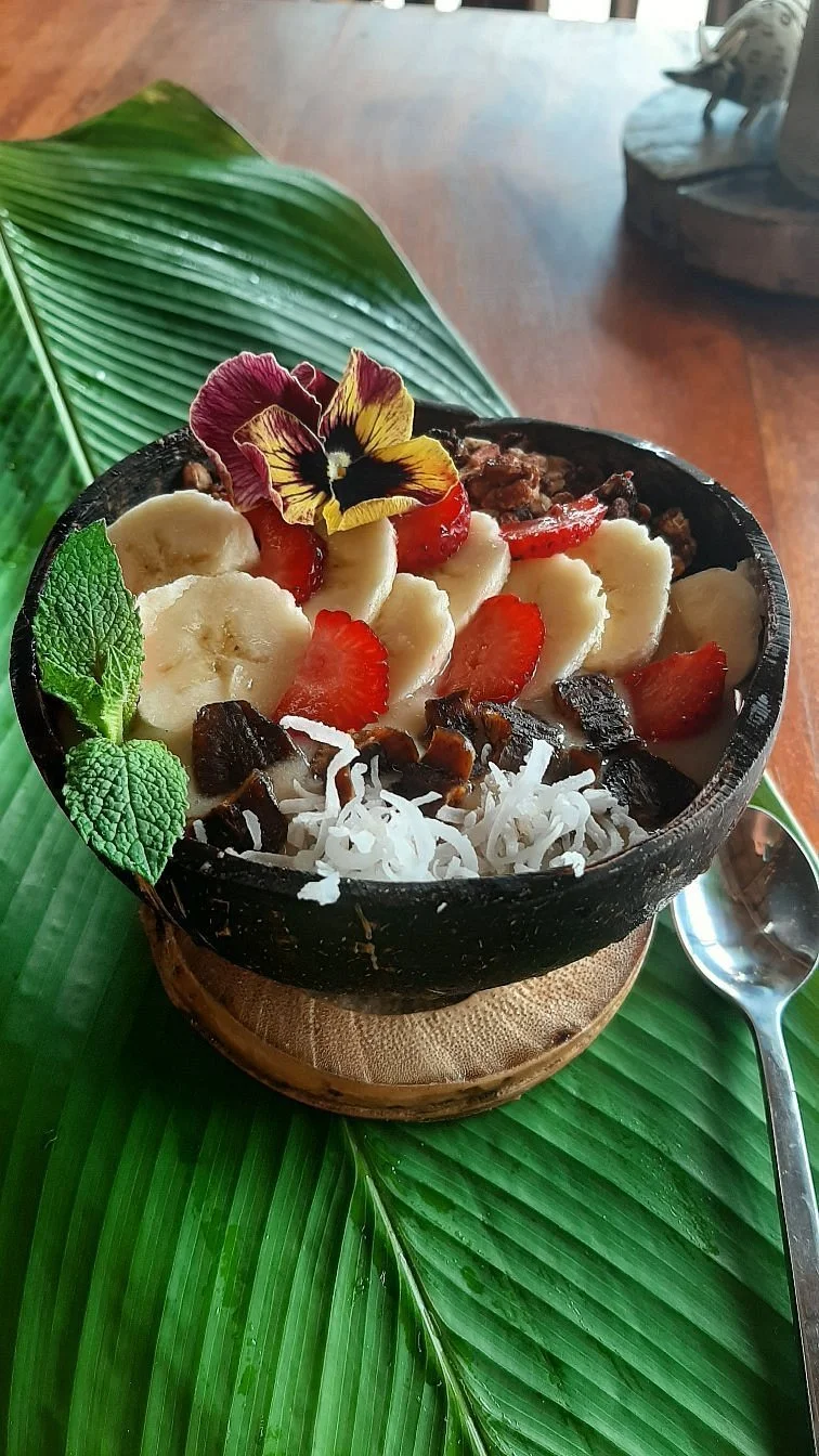 A smoothie bowl topped with sliced bananas, strawberries, shredded coconut, edible flowers, and mint leaves, served on a green leaf.