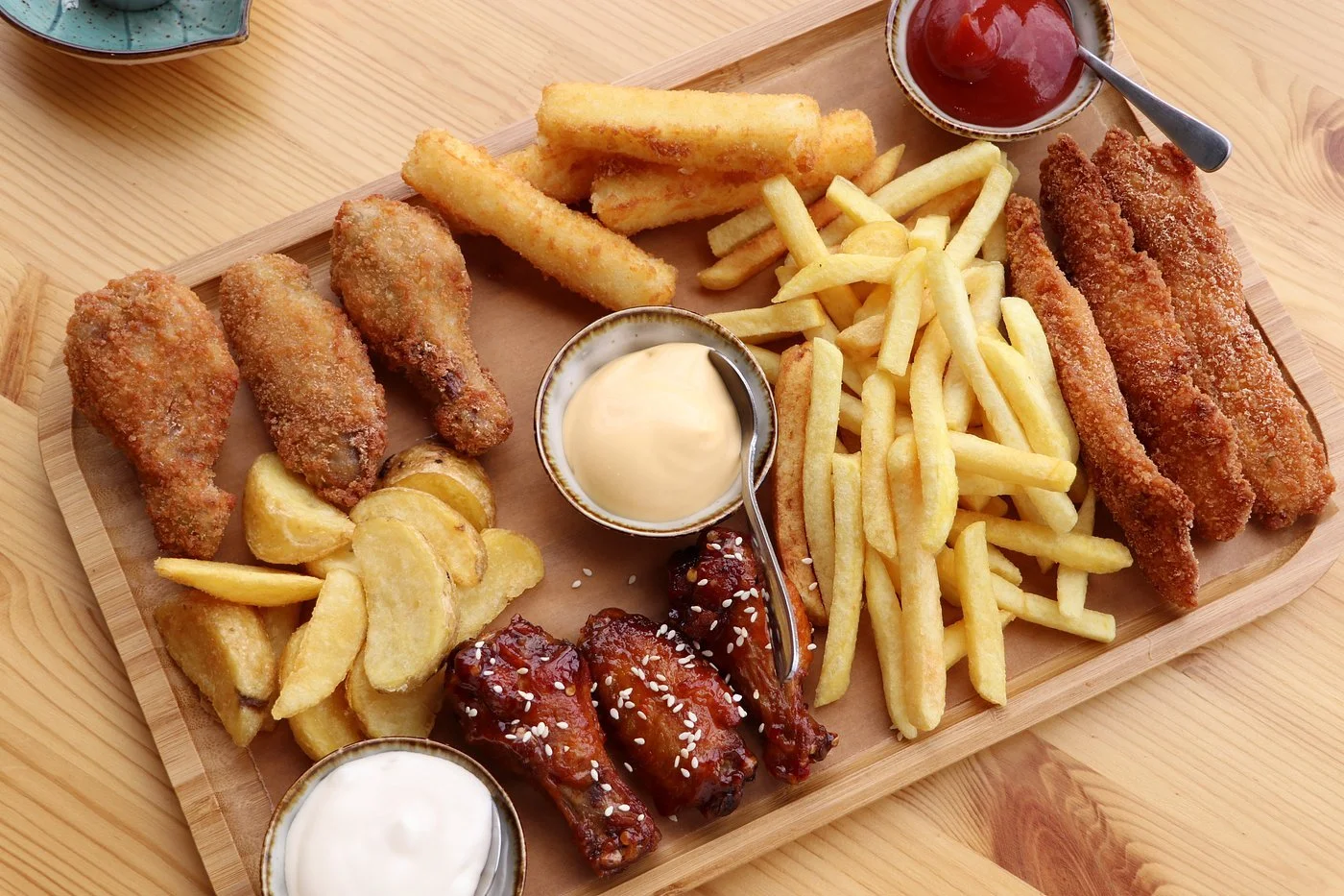 A wooden serving platter with fried chicken drumsticks, French fries, potato wedges, breaded fish fillets, and chicken wings topped with sesame seeds. Includes bowls of mayonnaise, ketchup, and ranch dressing.