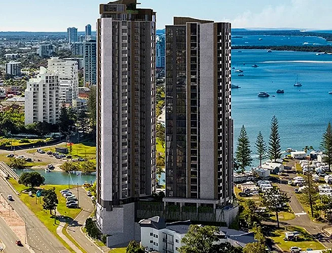 Two tall, modern high-rise buildings with many windows, located near a waterfront with boats and a park with trees in the background.