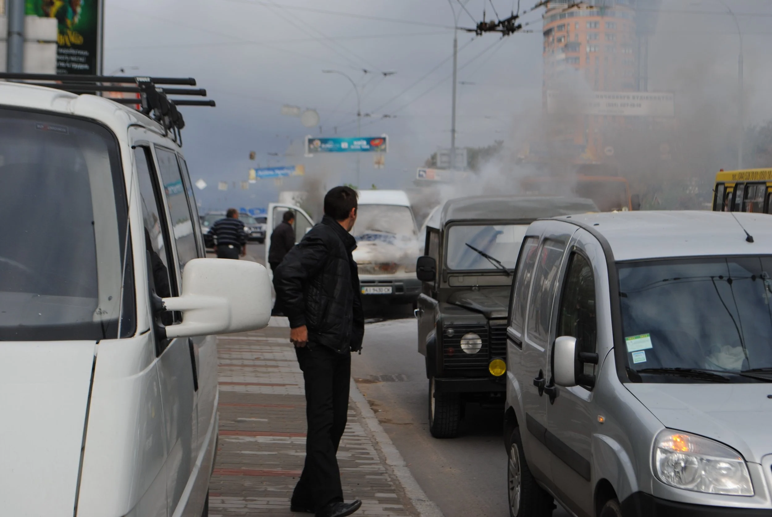 A street scene with several parked cars and people walking, shrouded in smoke or fog, with buildings and street signs in the background. Kiev November 2011, Marina Parrish ©