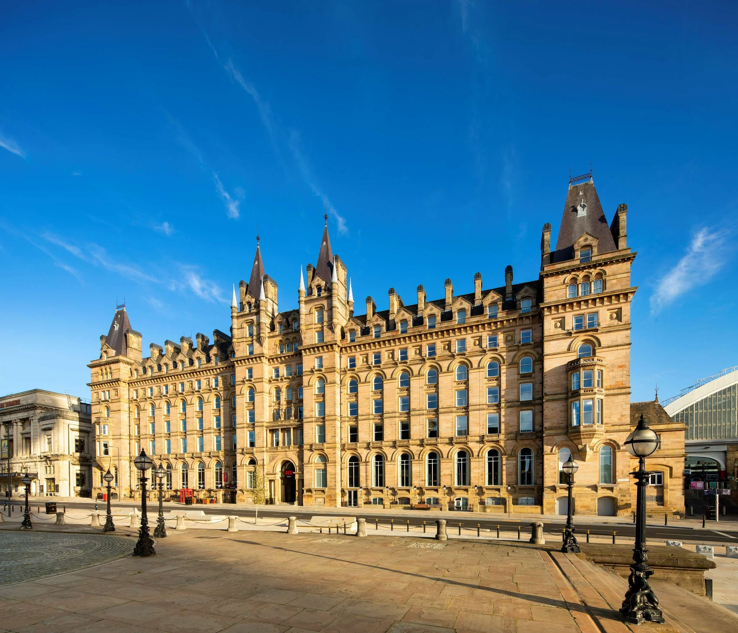 Historic building with multiple towers and turrets, ornate architecture, and numerous arched windows, set against a bright blue sky with a few wispy clouds, in an urban setting with street lamps and barricades in the foreground.
