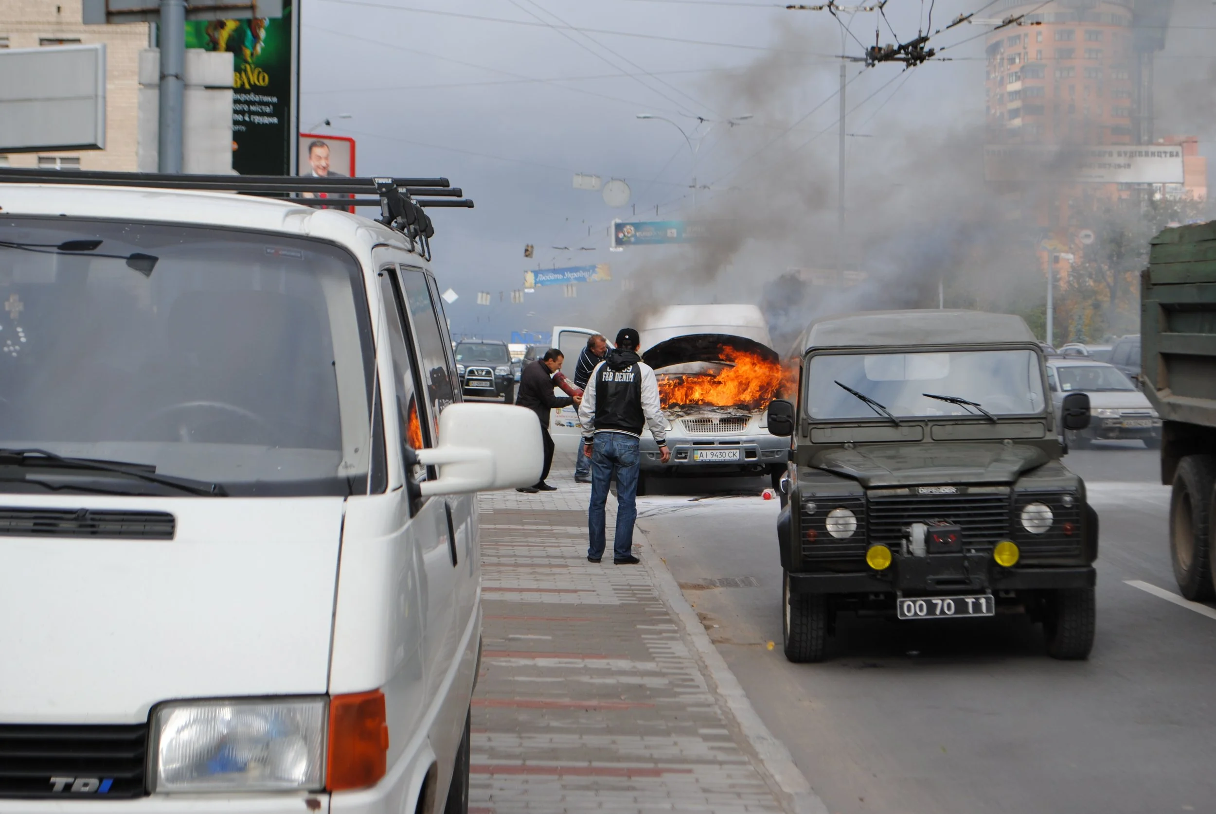 A car is on fire in the middle of a city street with thick black smoke, as people observe nearby. Vehicles are parked along the street, and buildings with billboards and signage are visible in the background. Kiev November 2011, Marina Parrish ©