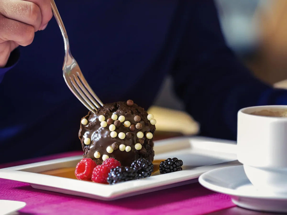 Person holding a fork over a chocolate dessert with white and chocolate sprinkles, served with raspberries and blackberries on a white plate, with a cup of coffee nearby.