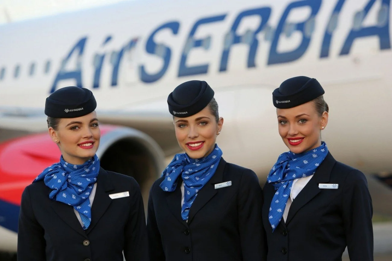 Three female airline crew members in uniform standing in front of an airplane, smiling at the camera.