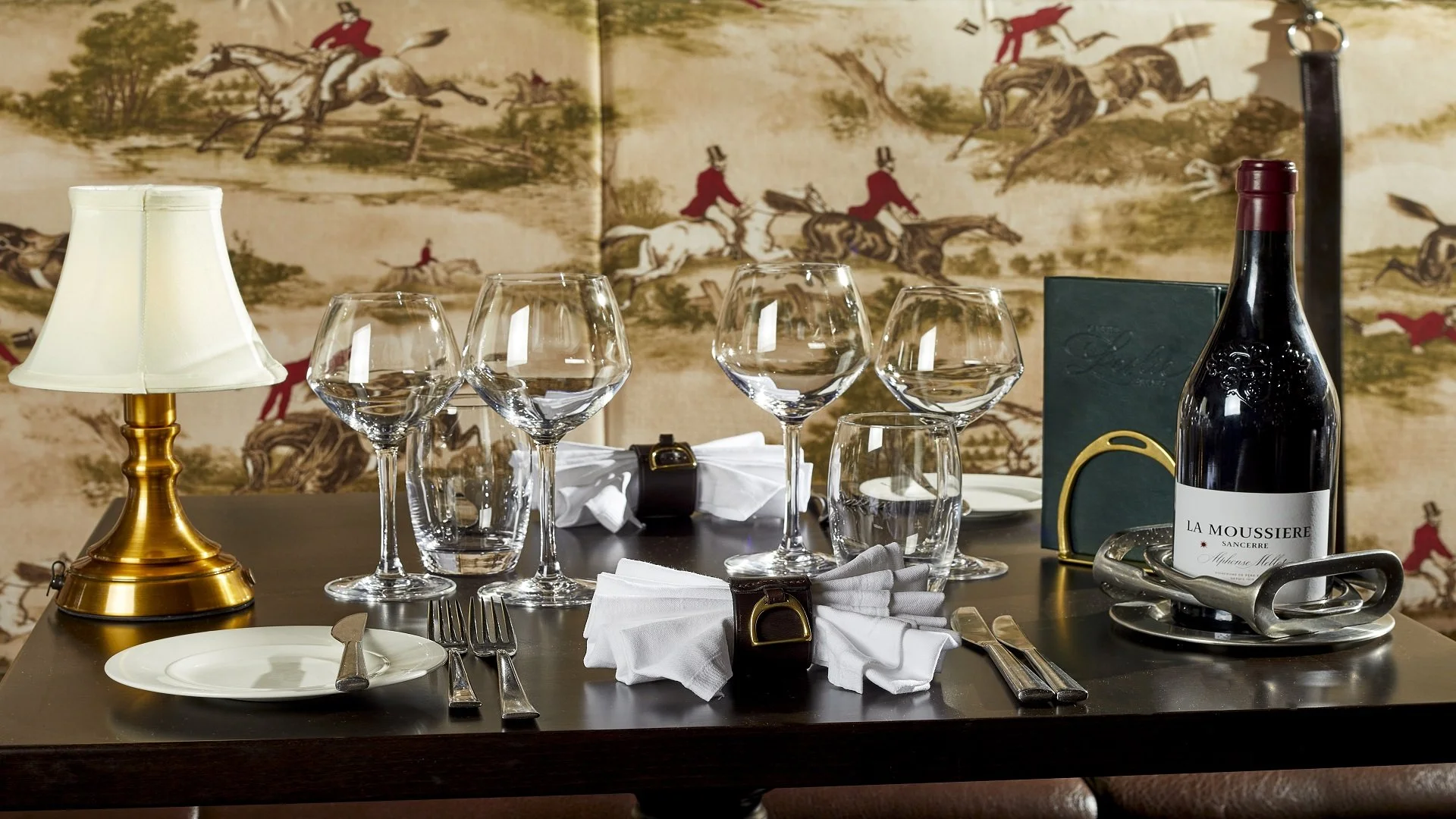 A dinner table setting with a gold lamp, multiple wine glasses, a bottle of red wine labeled 'La Moussiere', a green menu, and silverware with white napkins in front of an artistic landscape painting with horses and riders.