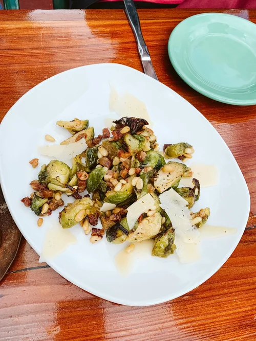 Plate of roasted Brussels sprouts with pine nuts, bacon bits, and shaved parmesan cheese on a white dish
