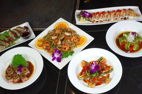 Assorted Asian dishes on white plates and bowls, including sushi rolls, shrimp with noodles, cucumber and meat rolls, and stuffed peppers, garnished with edible flowers and greens.
