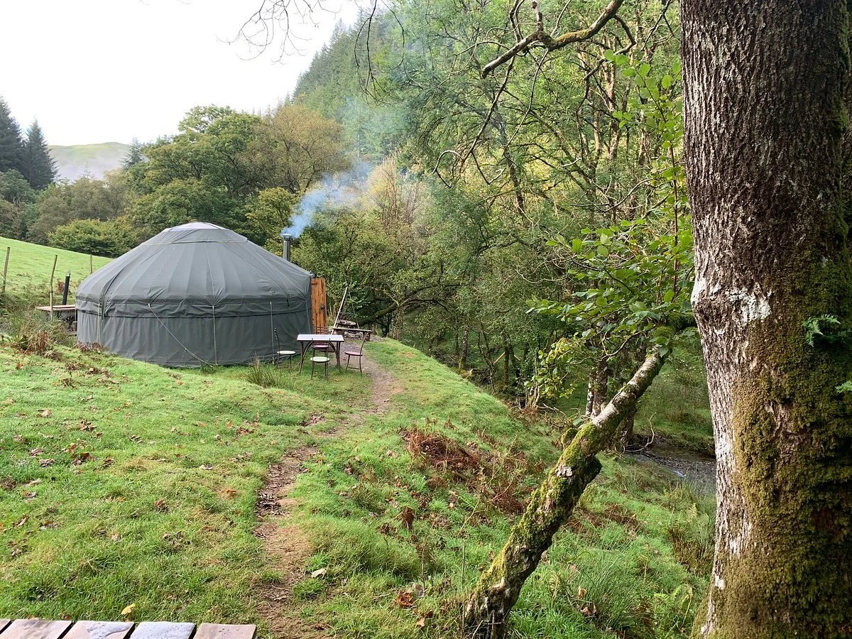 A portable yurt tent on a grassy hillside surrounded by trees, with a small outdoor table and chair, and a stream nearby in a forested area.