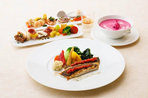 A plate with cooked fish, sautéed vegetables, and cauliflower, with a side salad, pink soup, and a small glass of drink in the background.