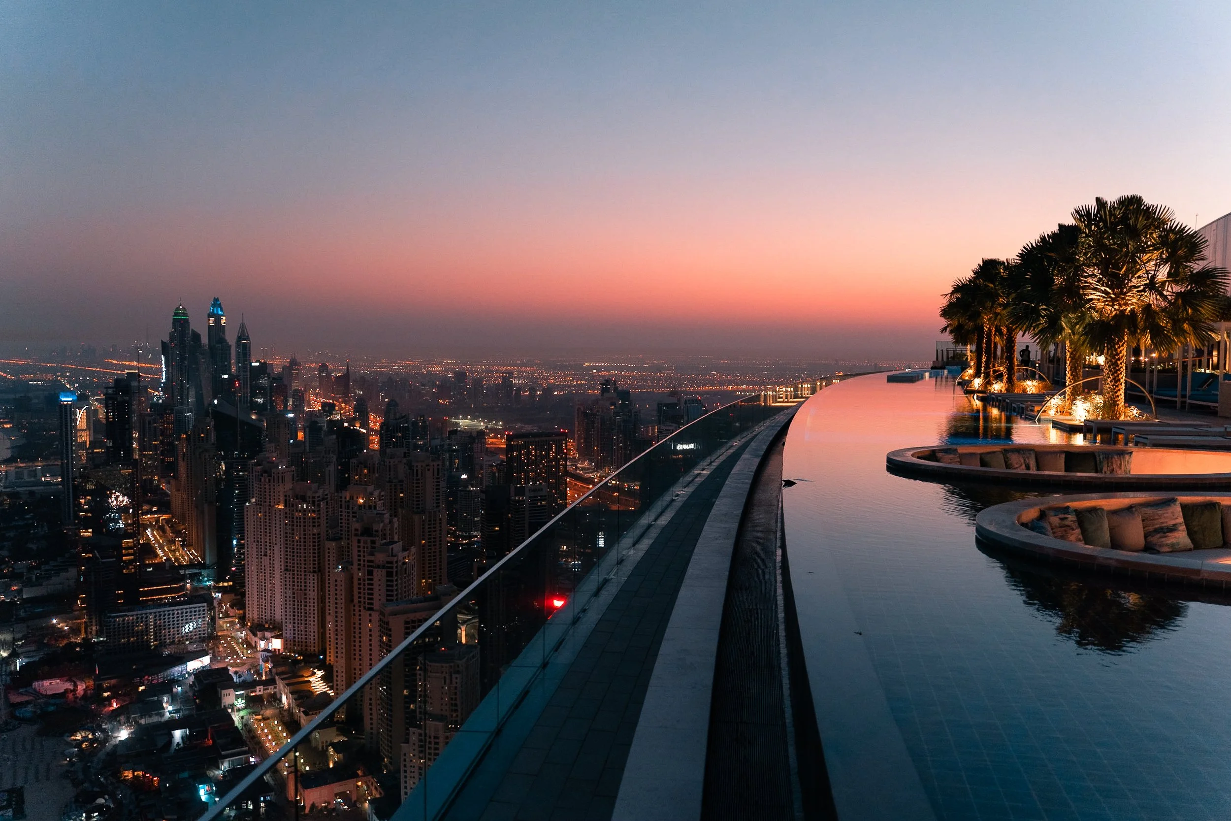 Nighttime view from a rooftop pool overlooking a city skyline with illuminated buildings, palm trees, and a colorful sunset sky.