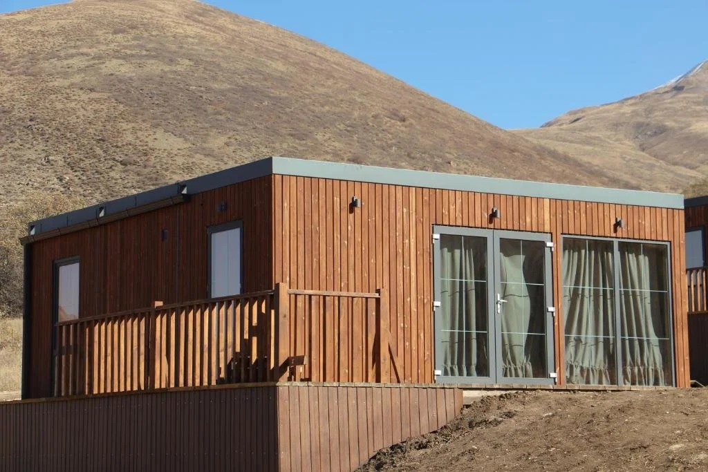 A modern wooden house with large glass sliding doors and curtains, set against a dry, mountainous landscape under a clear blue sky.