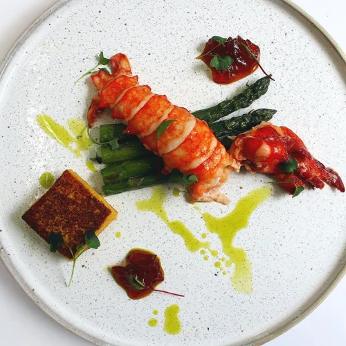 Seared lobster tail with grilled asparagus, a square of crispy polenta, and garnishes of tomato sauce and peppery microgreens, drizzled with green oil on a white plate.