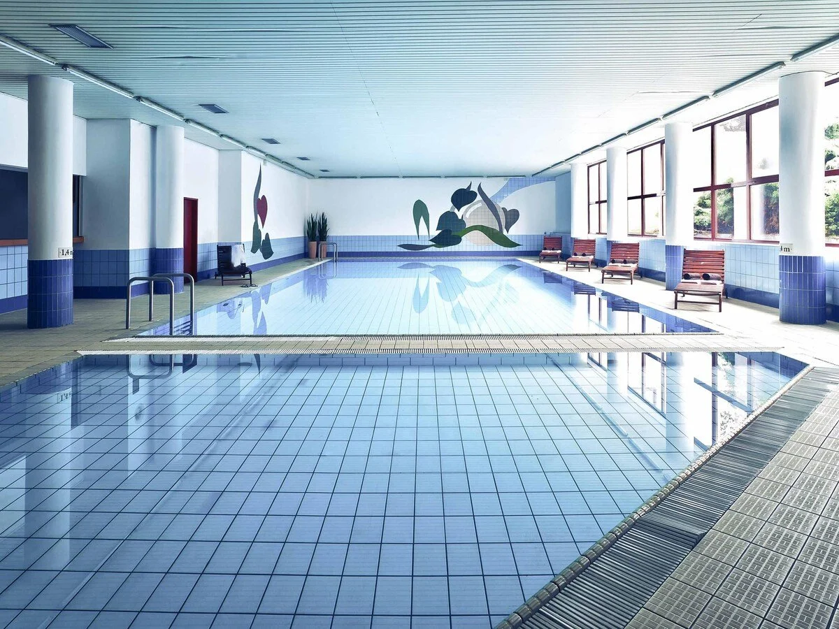 Empty indoor swimming pool with blue tiled floor, lounge chairs, and large windows letting in natural light.