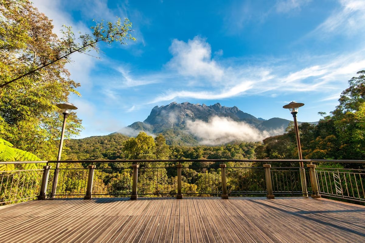 A wooden observation deck with a metal railing overlooks a lush forest and a mountain with clouds near its peak under a blue sky.