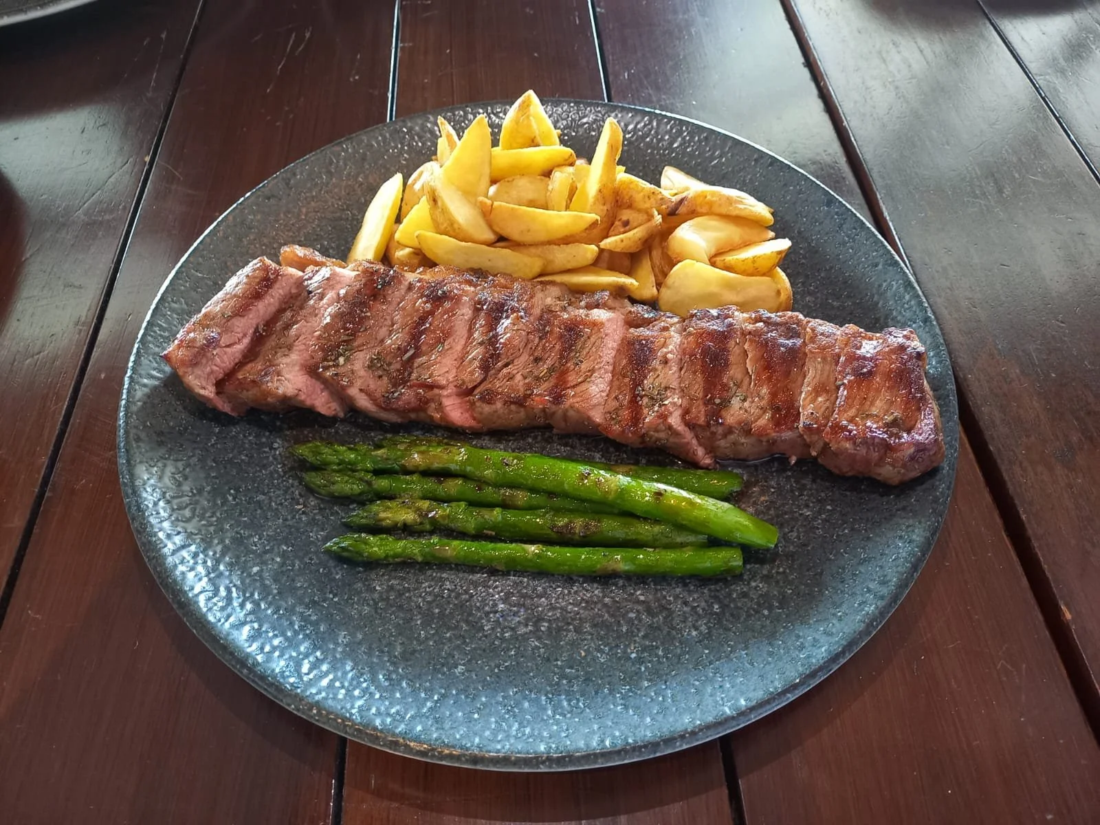 A plate of grilled steak with green asparagus, French fries, and potato wedges on a dark glass plate.