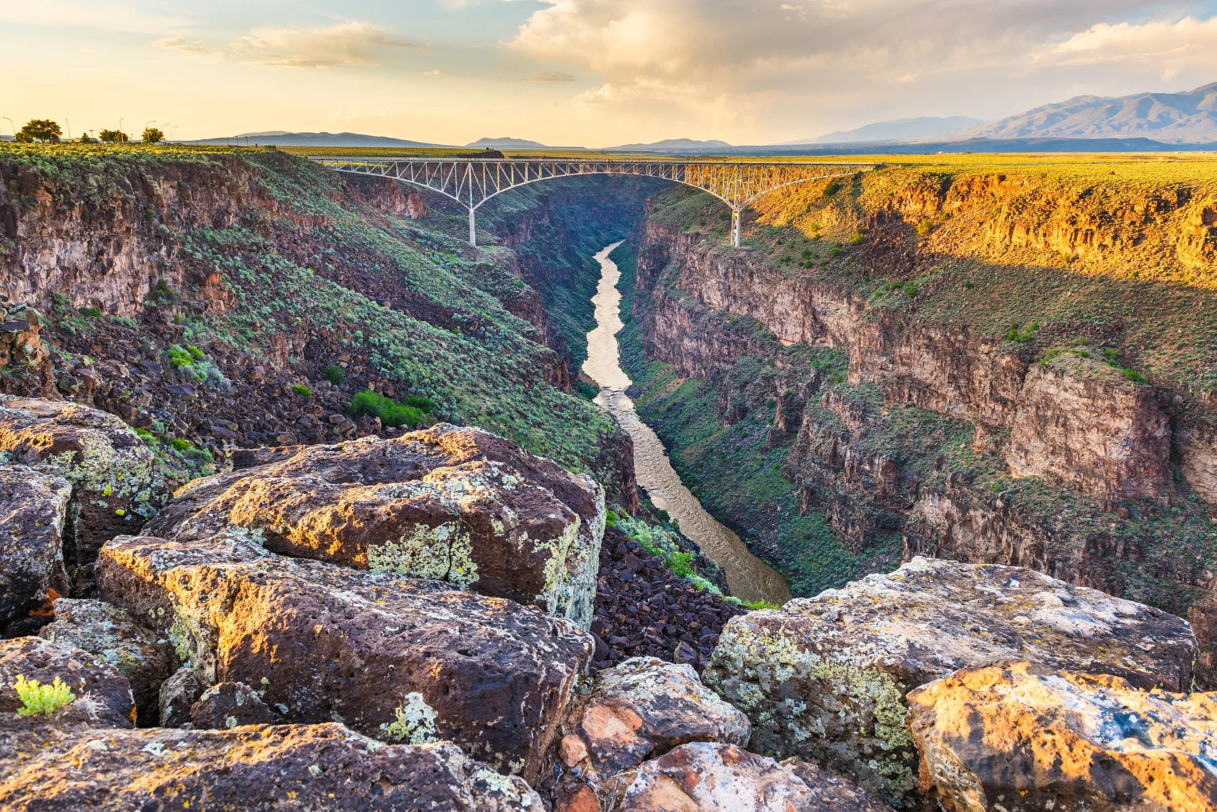 View of a canyon with a river at the bottom, a bridge spanning across the canyon, and mountains in the distance during sunset.