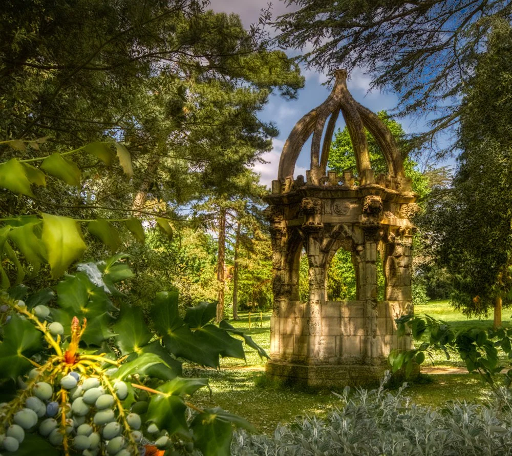 An ornate stone structure with Gothic architectural elements, surrounded by lush green trees and plants in a park.