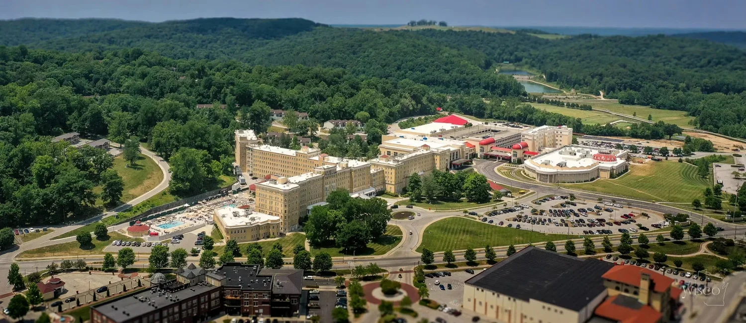 Modern mountain hotel balcony overlooking outdoor hot pools, steam rising, snowy landscape, and forested mountains in the background during dusk.
