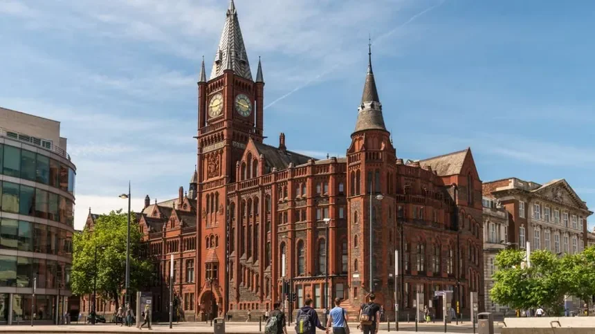 Red brick historic building with a clock tower and multiple turrets, situated on a city street with pedestrians and modern buildings nearby, under a blue sky.