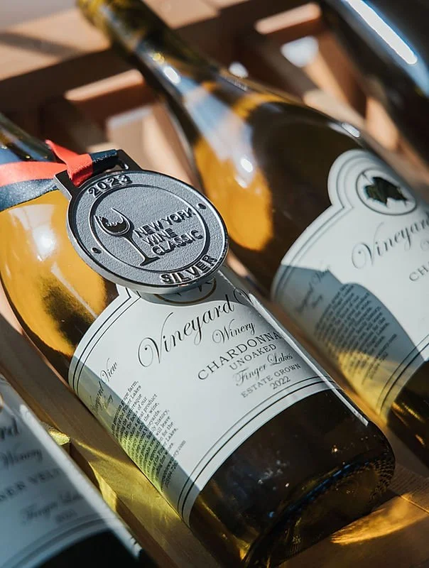 Two bottles of Chardonnay wine from Vineyard Winery resting on a wooden surface, with a silver medal award hanging from one of the bottles.