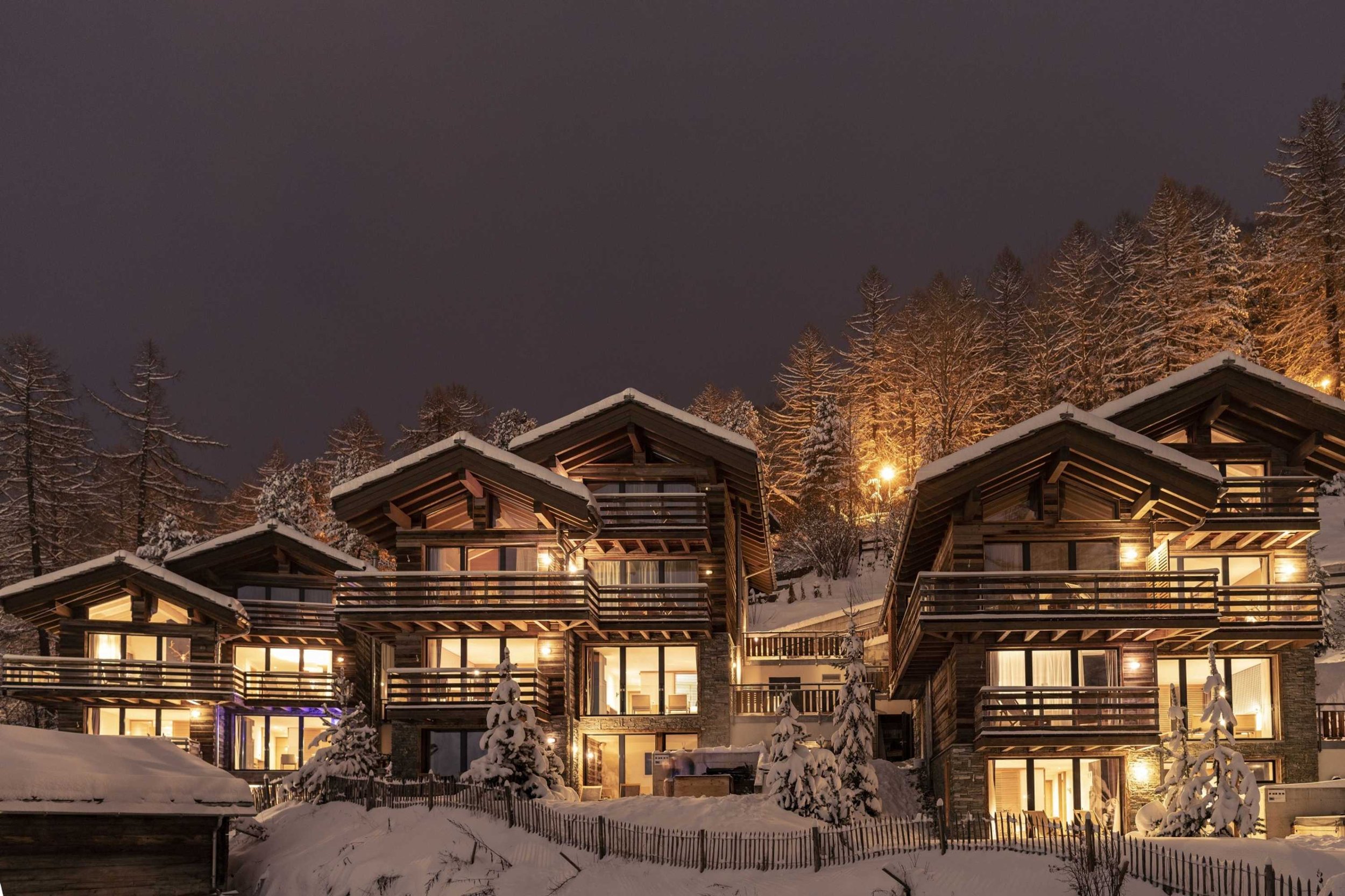 Nighttime view of snow-covered chalet-style cabins with illuminated windows, set against snow-covered trees and dark sky.
