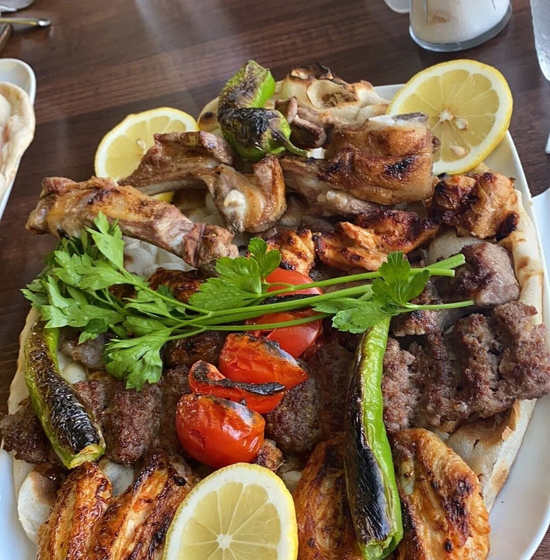 A plate of grilled meats including chicken, beef, and lamb, garnished with lemon slices, cherry tomatoes, charred green peppers, and fresh parsley.