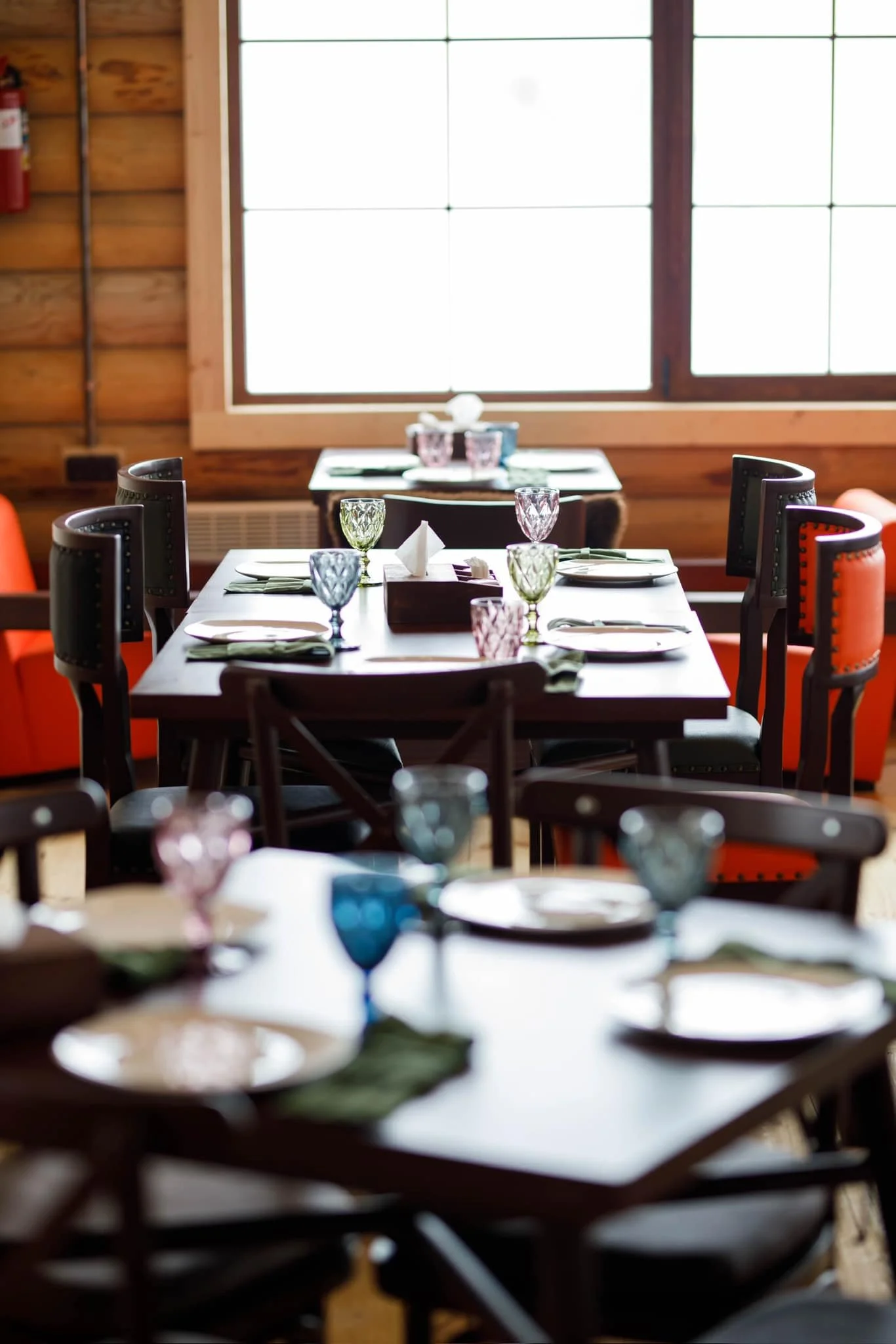 A dining area with a wooden interior, set for a meal with plates, colorful glassware, napkins, and cutlery on a dark wood table.
