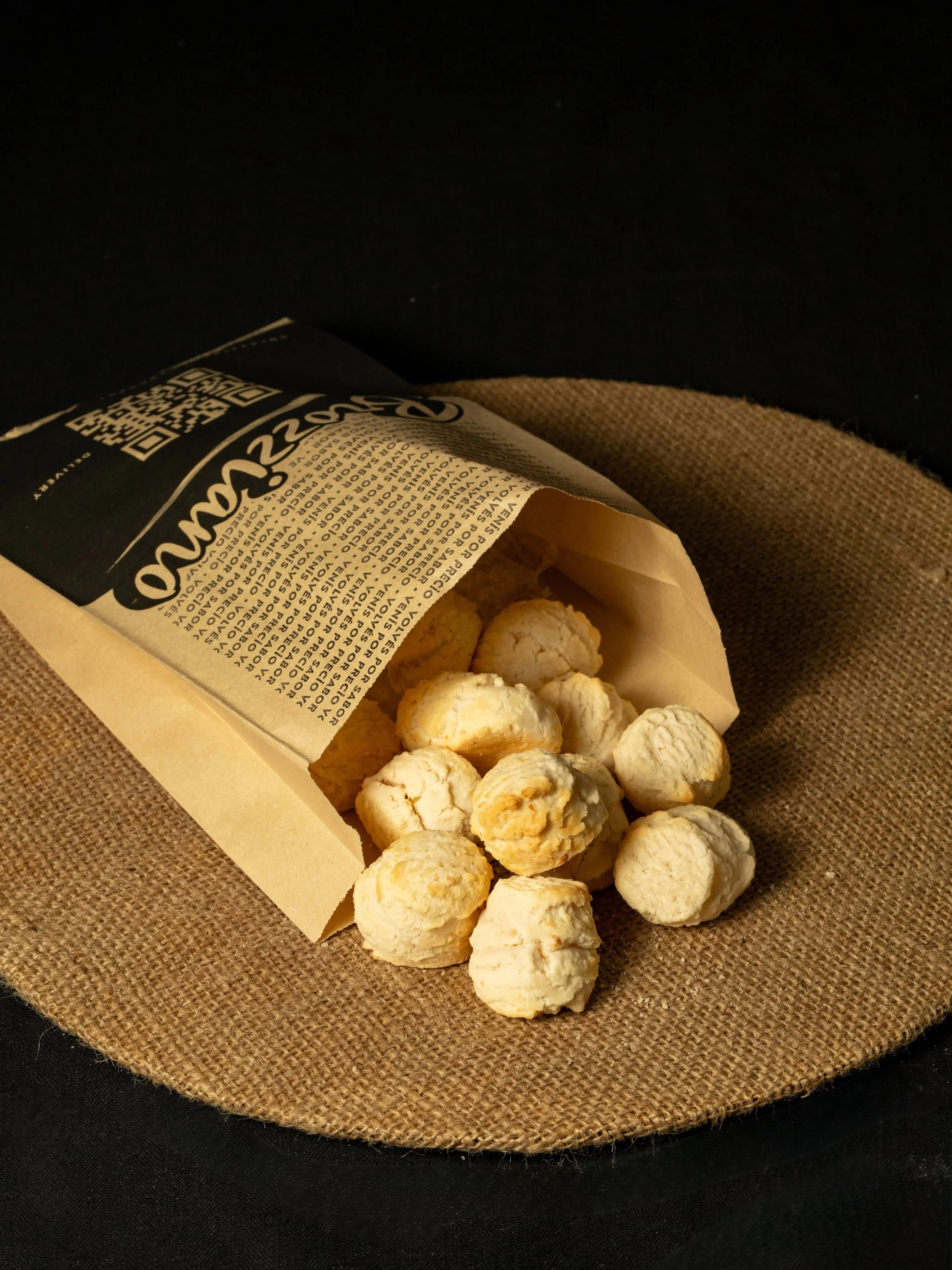 A paper bag of cheese curds resting on a round piece of burlap fabric, with a dark background.