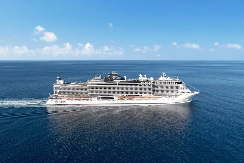 A large white cruise ship with yellow and black funnel sailing on calm blue ocean water with distant land in the background.