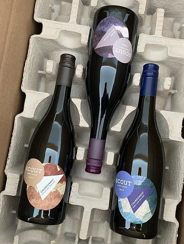 Three bottles of Scout Winery Chardonnay wine in a cardboard wine shipment tray, with a black bottle in the middle and two bottles on either side, one with a beige label and the other with a blue label.