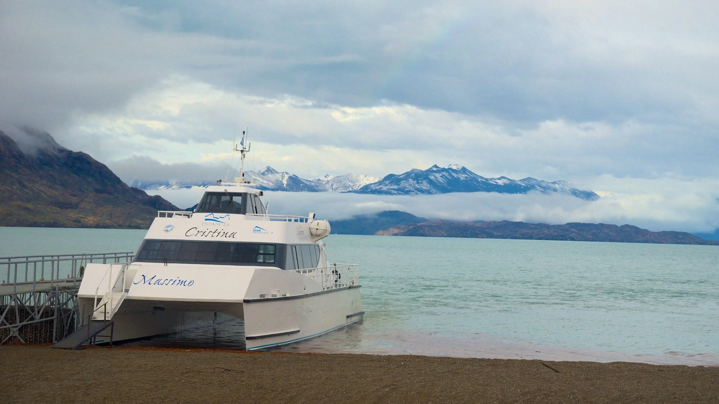 A white yacht named 'Massimo' docked at a pier on a body of water, with mountain ranges in the background under a cloudy sky.