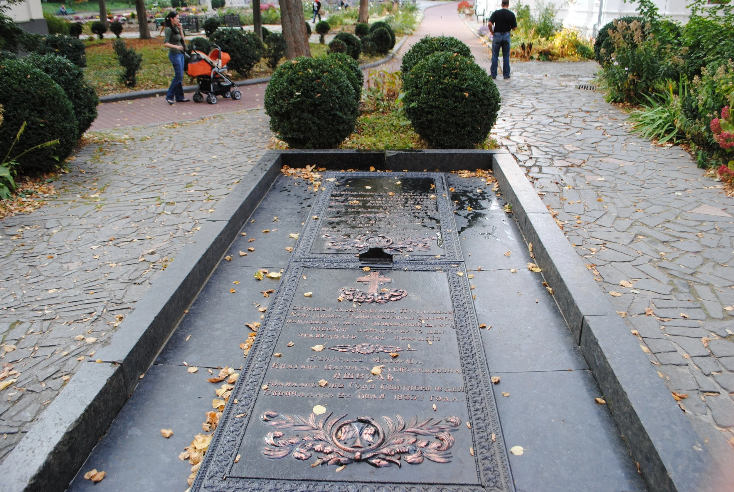 A park scene with well-maintained bushes, trees, and pathways. In the foreground, there is a large, decorative metal grave marker with Russian inscriptions, fallen leaves, and a small water puddle. In the background, two people are walking, one with 